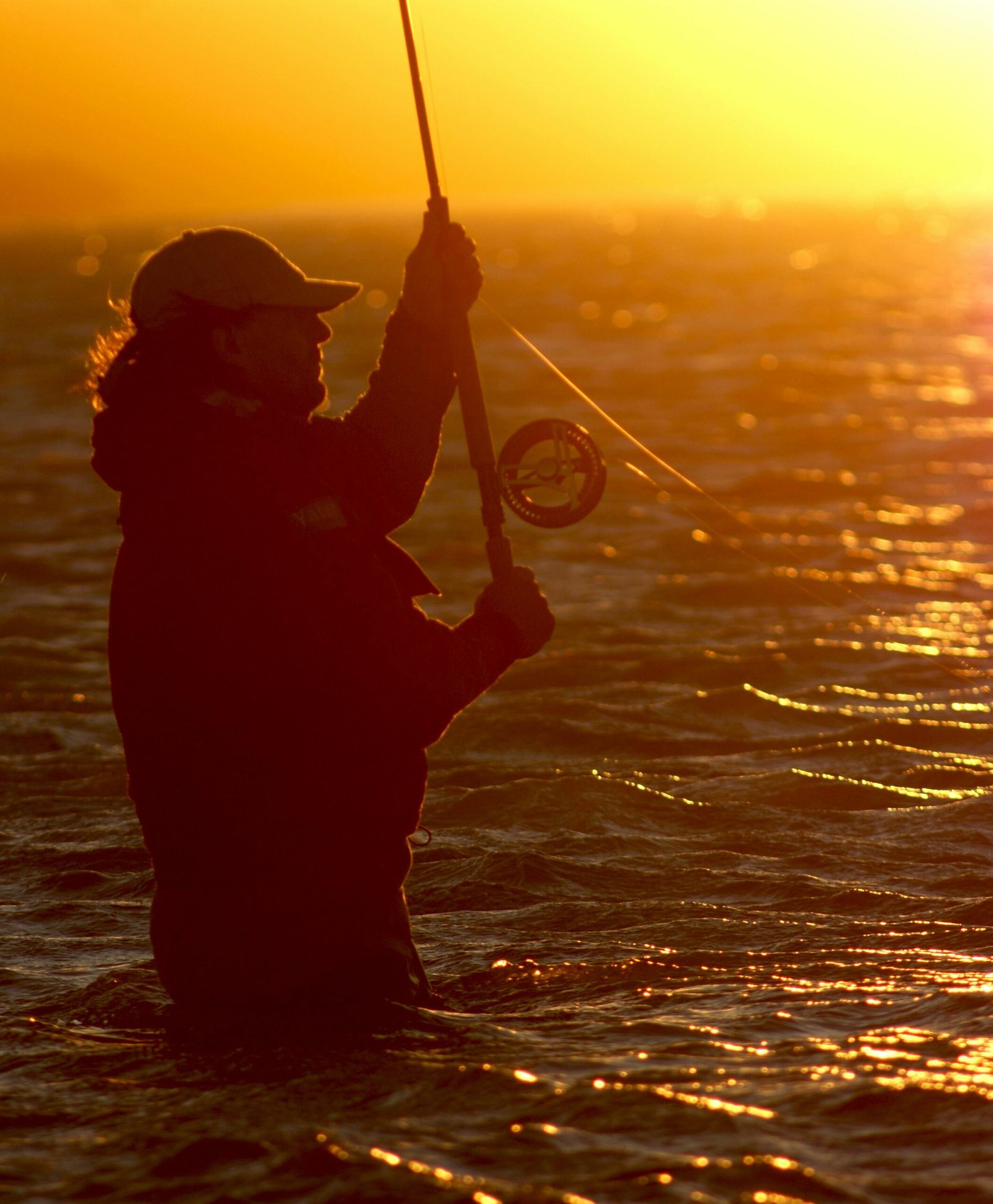 Silhouette of a man fishing at sunset, casting a fishing rod into the water with golden light reflecting.